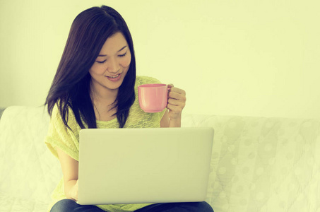 快乐的年轻亚洲妇女使用笔记本电脑和持有粉红色杯咖啡室内。复古风格