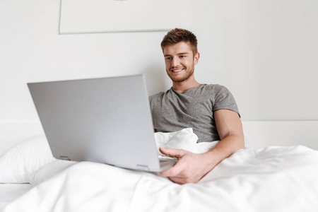 微笑的年轻人工作在笔记本电脑, 而坐在床上在家