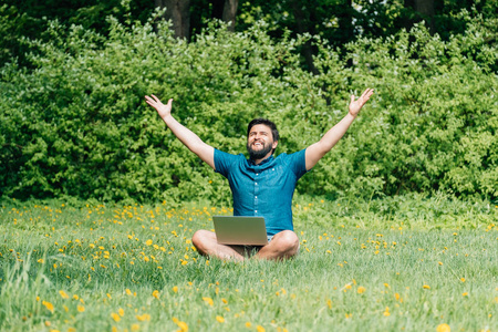 快乐兴奋的人用手提电脑举起手庆祝成功的工作或伟大的新闻坐在绿色的草地上