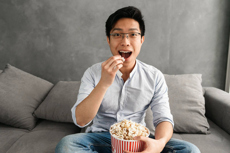 一个快乐的亚洲年轻人的肖像吃爆米花, 而坐在沙发上, 在家里看电视