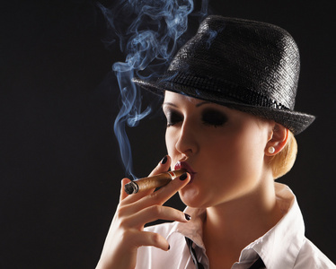 年轻有吸引力的女流氓吸烟雪茄在黑暗的背景照片