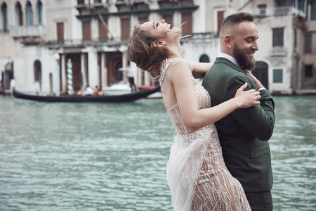 威尼斯运河附近的一对令人惊叹的新婚夫妇。穿着豪华象牙礼服, 凌乱的发髻头发, 穿绿色三件西装的男人笑女人