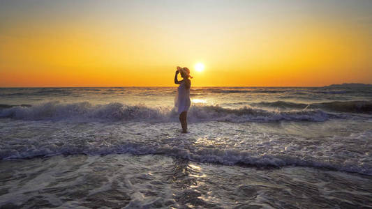 在日落背景下站在海岸线上的白色礼服和帽子的妇女