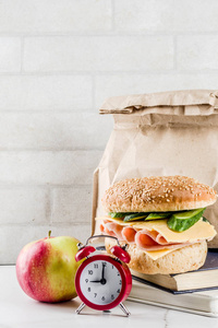 健康的学校食物概念, 纸袋与午餐, 苹果, 三明治, 书和闹钟在白色厨房桌拷贝空间