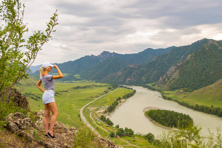 一个年轻的金发女郎穿着白色的帽子, 蓝色牛仔短裤和条纹 t恤矗立在岩石上, 朝阿尔泰山脉的 Katun 河望去。