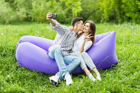 年轻情侣接吻坐在充气沙发 lamzac, 并采取自拍在电话上休息时, 在