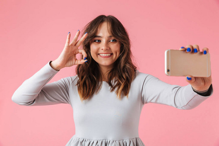 一个快乐的年轻女孩的肖像采取自拍手机和显示 ok 手势隔离粉红色背景
