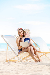 快乐的年轻母亲和孩子在海滩上的泳装使用平板电脑, 而坐在沙滩椅