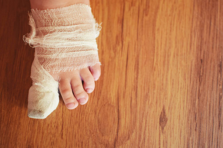 受伤痛苦左脚的小男孩的孩子与白色纱布绷带拇指上的棕色地板背景顶部