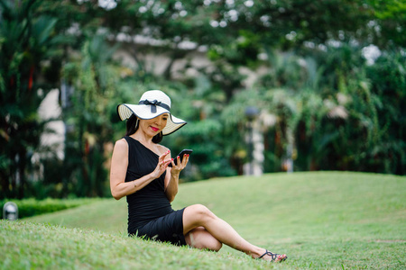 一个时尚的亚洲妇女在黑色礼服和白色帽子公园与智能手机的肖像