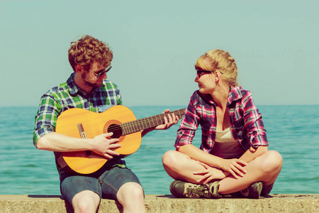 年轻男子玩吉他给他的女朋友室外阳光灿烂的夏天一天由海边约会浪漫的情侣