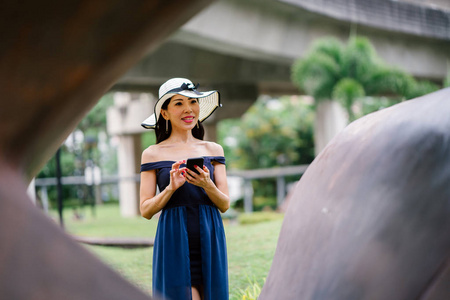 一个时尚的亚洲妇女的肖像在蓝色的礼服和白色的帽子公园与智能手机