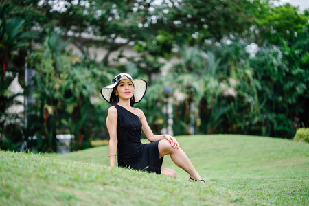 亚洲华人妇女肖像在一个优雅的黑色礼服摆在一个白色宽边的帽子, 白天在公园里