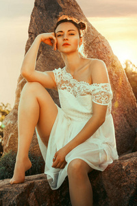 美丽的夏天户外肖像的年轻女子在白色礼服在日落灯在公园。坐在石头上, 享受自然, 温暖的色调