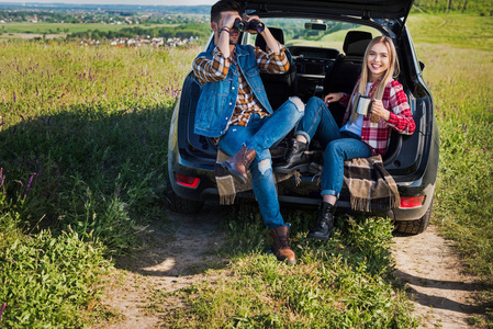 男性游客看着望远镜, 而他的微笑的女朋友坐在附近的汽车树干在农村领域的咖啡杯