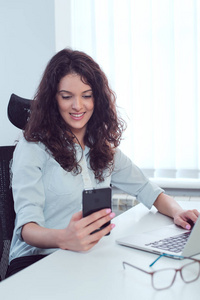 年轻的女商人坐在她的办公桌在现代办公室检查手机上的信息, 并在笔记本电脑上工作