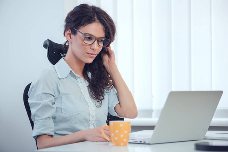 女商人戴眼镜, 在办公桌边喝咖啡, 看笔记本电脑屏幕, 在办公室微笑。