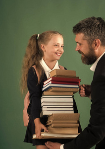 教育和回学校概念。孩子她爸给堆书。父亲和女生带着笑脸上深绿色的背景。女孩的马尾辫和校服与留胡子的人