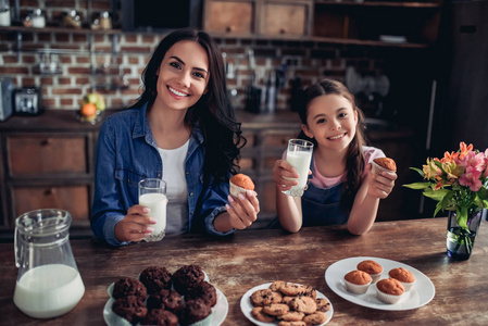 微笑的女儿和母亲的肖像拿着杯子牛奶和蛋糕在手中, 看着厨房里的相机