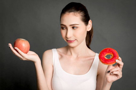 在红色苹果和卡路里甜甜圈之间的妇女举行和选择灰色背景。健康饮食与垃圾食品概念