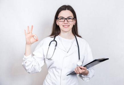 一个年轻的微笑的女人黑发医生在眼镜和医疗白大衣与听诊器和文件文件夹在白色背景