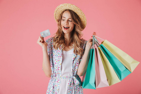 一个年轻快乐的女人在夏天的礼服和草帽看塑料信用卡, 而持有购物袋, 在粉红色背景隔离