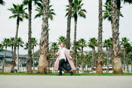 时尚快乐的年轻女子穿着粉红色的大衣走在公园旁边的绿色棕榈和享受天气