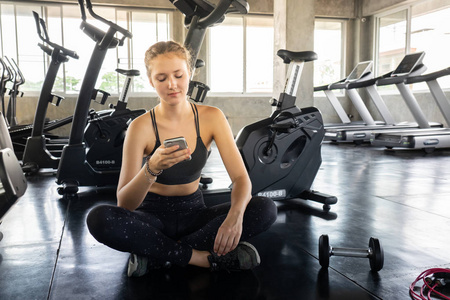 活跃的女孩使用智能手机在健身馆。年轻的微笑的妇女在健身房放松和听音乐使用手机