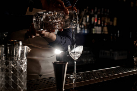 女酒保在白色围裙倾吐透明酒精饮料从测量杯子入鸡尾酒玻璃在酒吧柜台