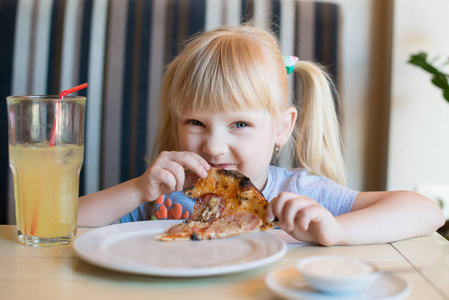 一个小快乐的女孩坐在桌子上的咖啡馆和吃新鲜的比萨饼与香肠。她长着金黄色的头发。