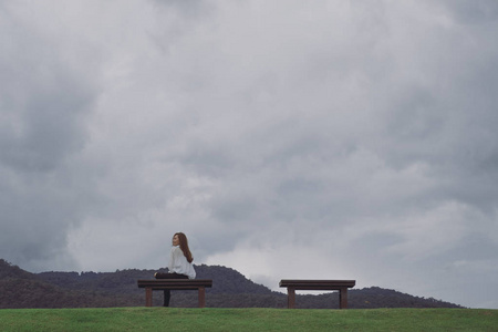 一个快乐的女人坐在公园的木凳上, 多云, 阴沉的天空背景照片