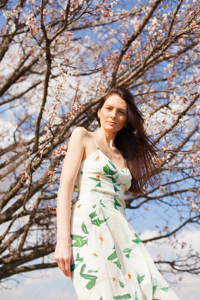 在灿烂的阳光下在盛开的花园里有着蓝眼睛的修长棕色头发的女人