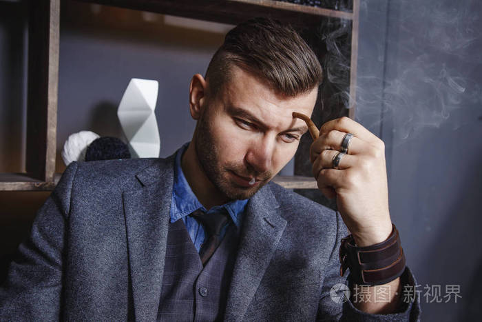 时尚英俊的野蛮男子在商业灰色西装和 hairdress 坐在椅子和吸烟管道.