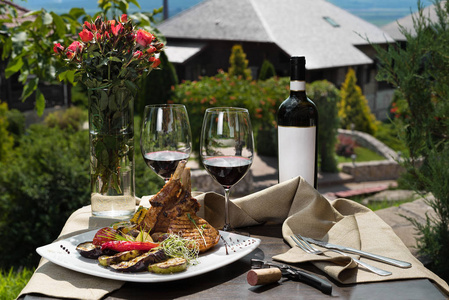 浪漫晚餐在外面.一盘猪肉排骨和两杯红酒, 绿草和花的背景照片