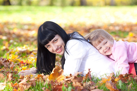 美丽的年轻母亲和她的女儿躺在秋天树叶上