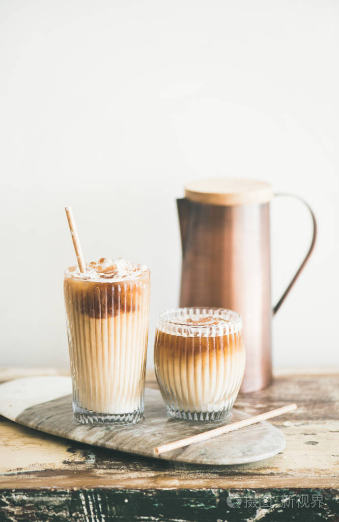 冰咖啡在高杯子与牛奶和秸杆在板上在质朴的木桌, 白色墙壁和水罐在背景上