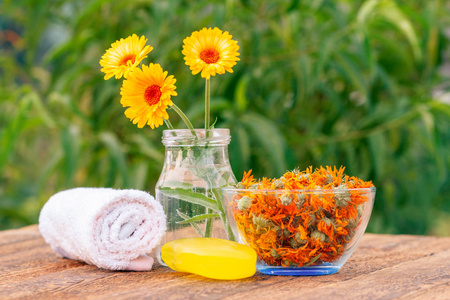毛巾, 肥皂与万寿菊提取物, 金盏花花卉在一个玻璃罐子和碗与干燥的花的金盏花在木板上绿色自然背景。水疗产品及配件