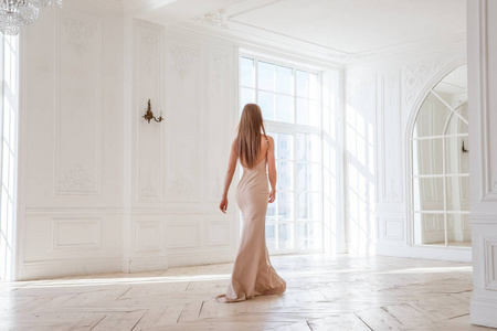 时装拍摄的美丽的金发女子在米色礼服站立反对白色内部背景