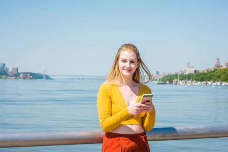 美国十几岁的女孩在纽约旅行, 穿着黄色长袖深 v 颈部适合作物顶部, 红色橙色裤子, 站在哈德森河, 手机短信。桥梁, 海港在远