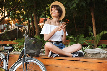 微笑的年轻女孩在夏天衣裳坐在公园用自行车和使用移动电话