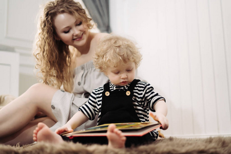 快乐卷曲的年轻母亲坐在地板上与她的小儿子, 读一本书, 并有乐趣