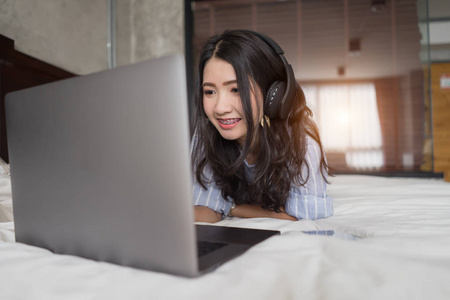 女人在床上使用笔记本电脑听音乐。从笔记本电脑收听音乐的年轻亚洲女孩蓝牙头电话