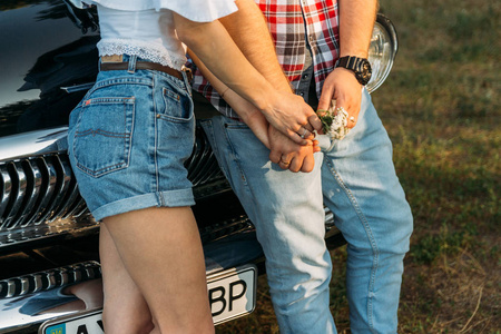 一个男人和一个女人牵着手的躯干, 手上拿着一朵花和一只钟。站在车边检查衬衫, 短裤和牛仔裤, 白色上衣