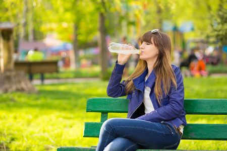 美丽的时尚女人与手提包坐在公园里, 等待在长凳上喝水