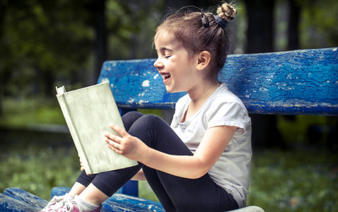 小可爱的女孩坐在长椅上, 在公园里读一本关于绿色自然背景的书。
