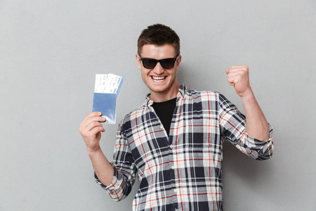 一个兴奋的年轻人在太阳镜的肖像持有护照门票和庆祝灰色背景