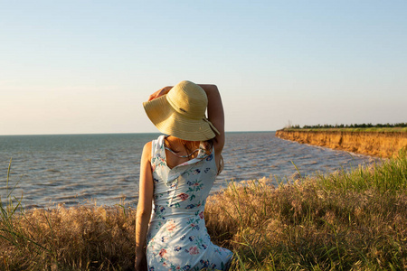 年轻的浪漫妇女穿着蓝色的礼服和帽子在背景海日落。坐回相机。一个红头发的女孩。夏季时间