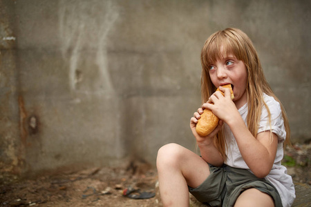 饥饿的无家可归的女孩吃一块面包在肮脏的小巷里