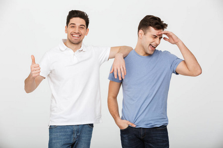 两个英俊的男人的朋友30s 穿着休闲 t恤和牛仔裤微笑着和摆在一起在相机与微笑拇指上孤立的白色背景
