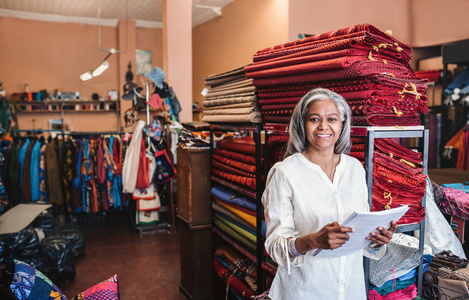 一个微笑成熟的面料店老板的肖像阅读文书工作, 而站在货架上充满五颜六色的布料和纺织品